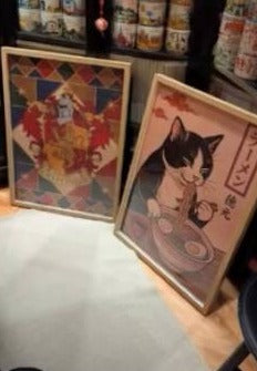 Dva uramljena japanska postera naslonjena na zid, među njima mačka sa ramenom